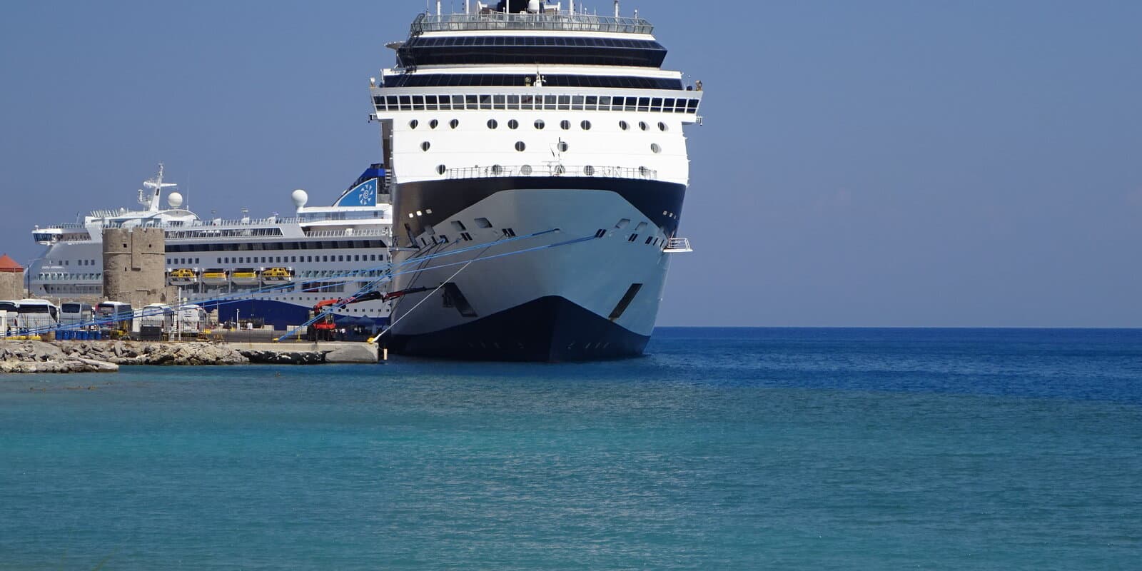 Celestyal Discovery and Celebrity Infinity moored at the quay in Port of Rhodes, Greece, with turquoise harbor water and a stone pier in the foreground