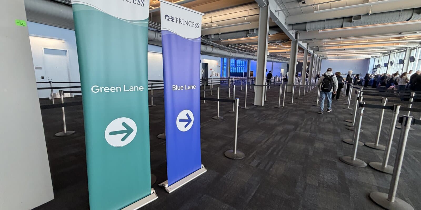 Travelers in a cruise terminal embarkation hall with lane signage before boarding