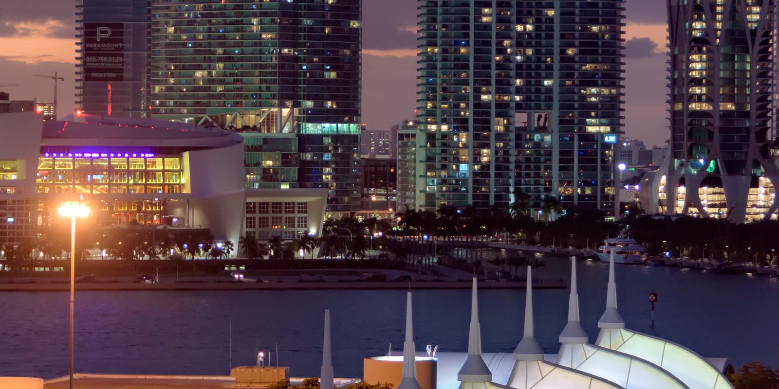 Illuminated sail-style roofs of the Miami cruise terminal at dusk with downtown high-rises beyond the harbor