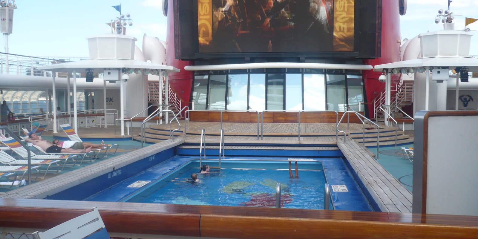 Families in a cruise ship pool on a sunny deck, with lounge chairs, a large outdoor movie screen, and a red funnel in the background