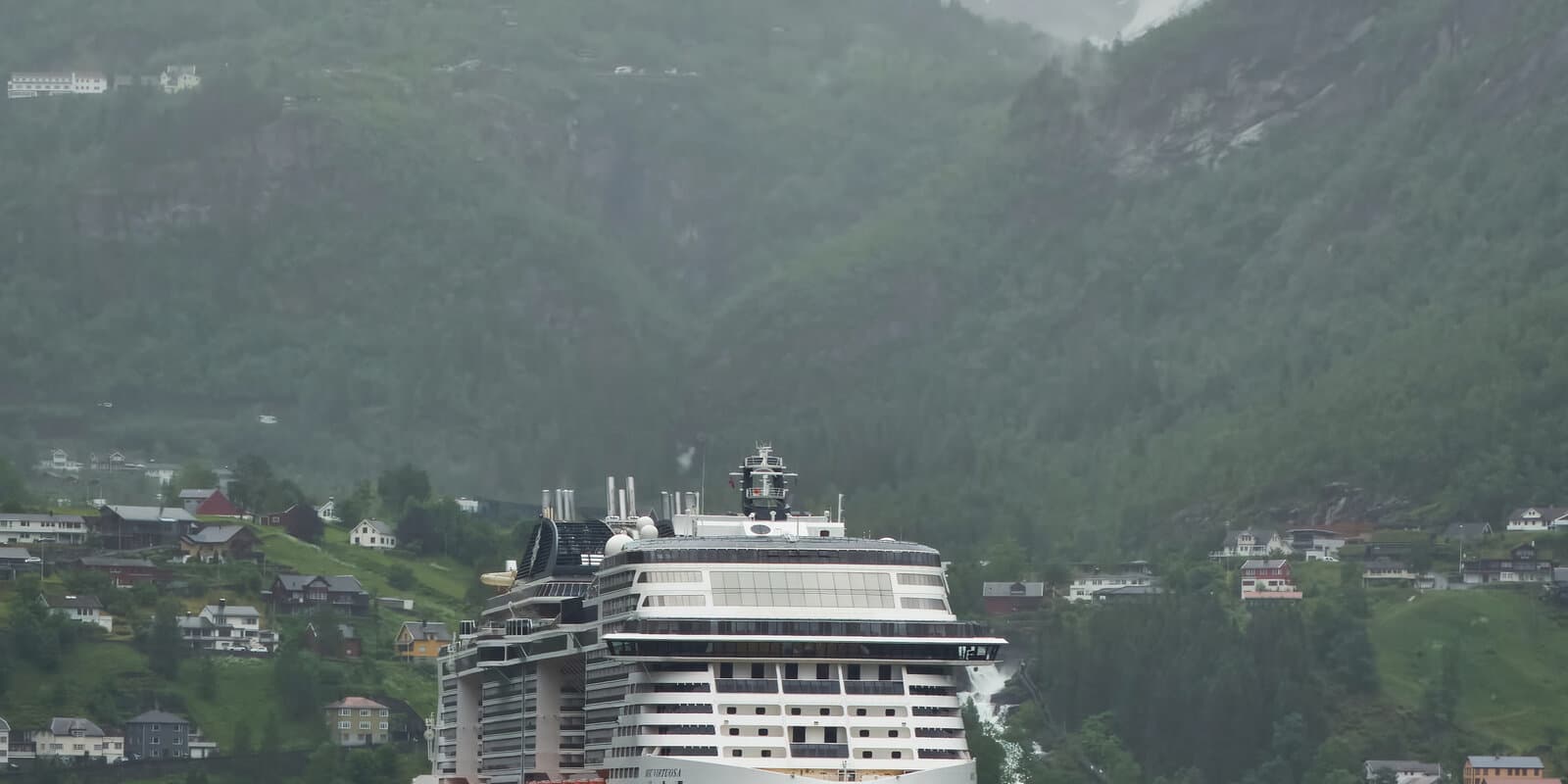 MSC Virtuosa cruise ship in profile at a Norwegian fjord pier, with MSC funnel colors and Geiranger mountains in the background