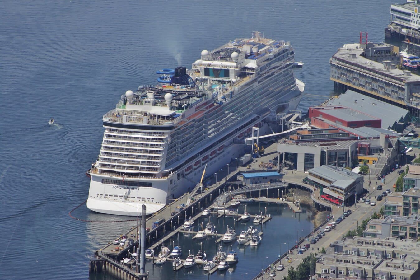 Norwegian Bliss docked at the Bell Street Cruise Terminal (Pier 66) in Seattle, with a marina beside the ship and downtown streets visible near the terminal building