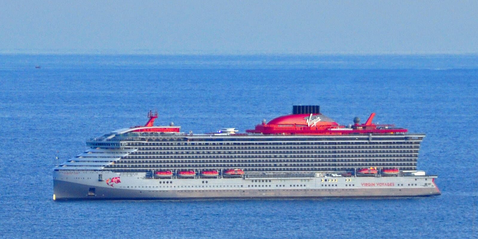 Virgin Voyages Scarlet Lady cruise ship departing Genova harbor with the city skyline behind the hull