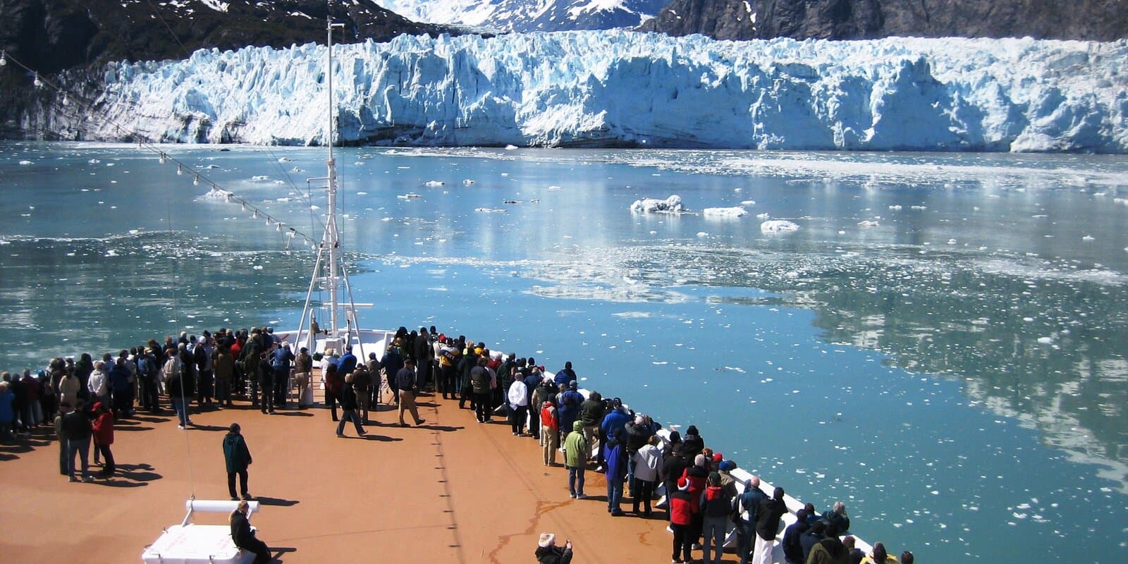 Passengers in layered jackets on a cruise ship deck viewing a blue tidewater glacier in Glacier Bay, Alaska