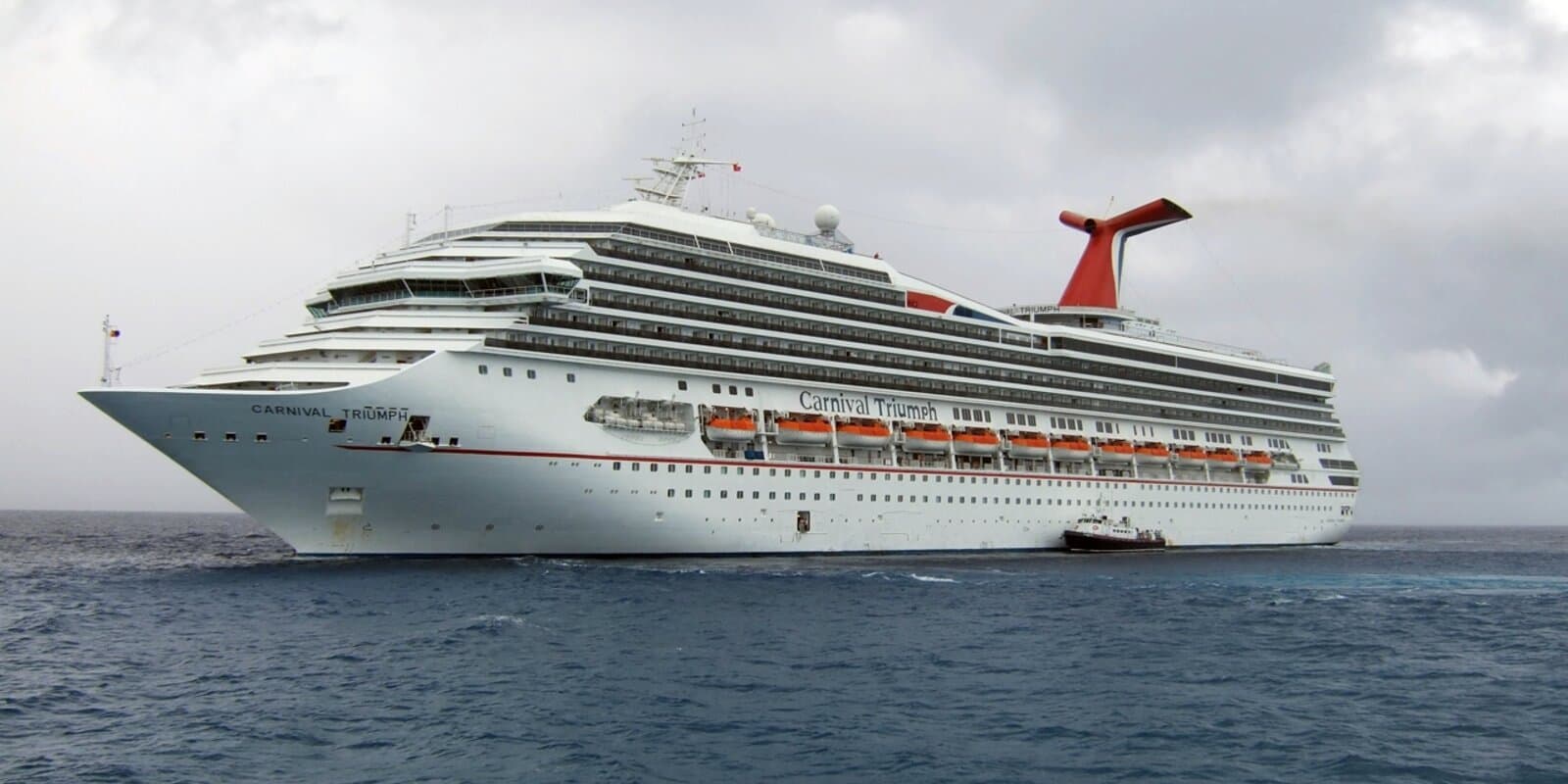 Carnival Triumph with red funnel on blue water, a tender boat alongside the hull