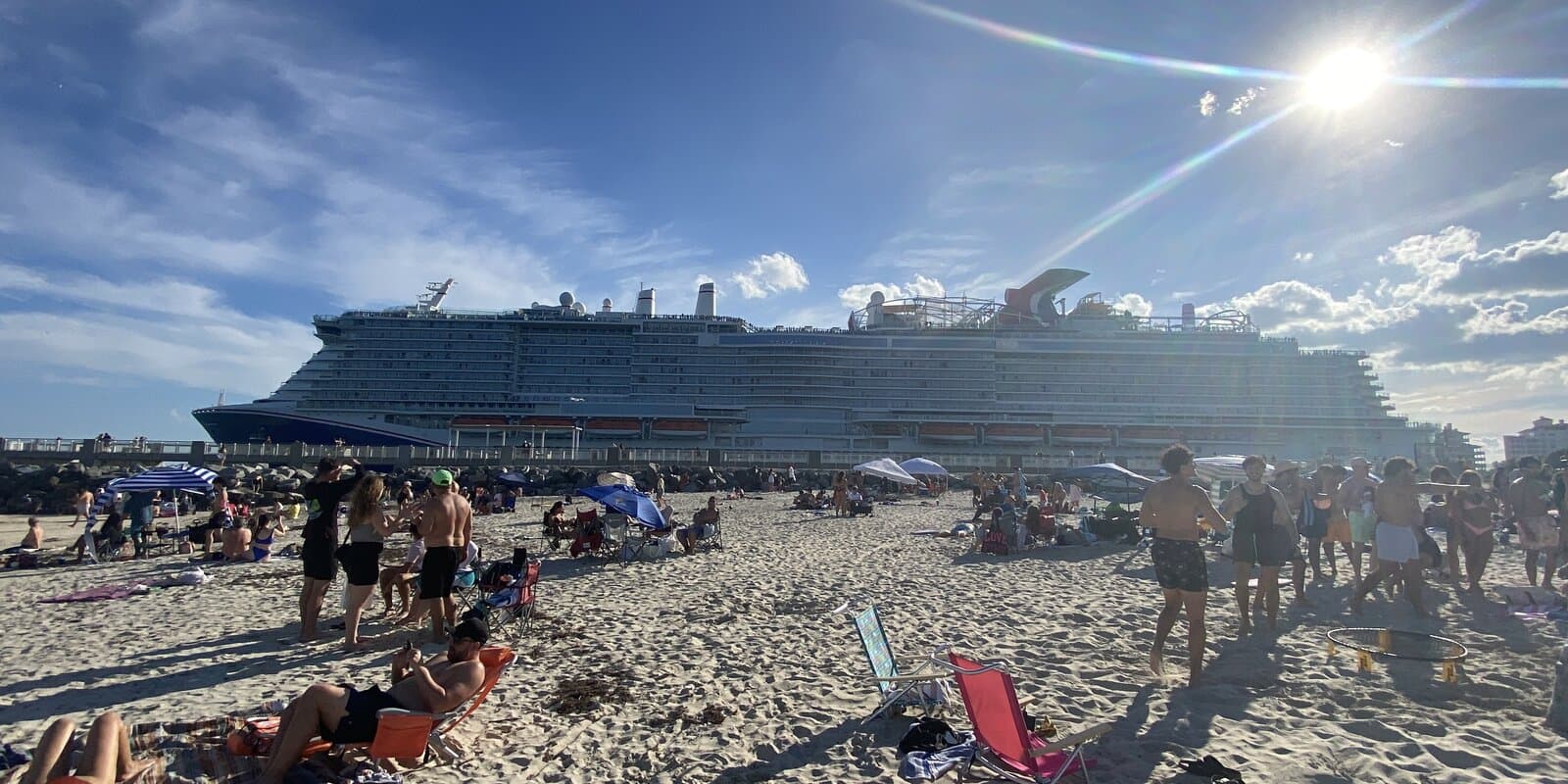 Carnival Celebration cruise ship docked at a pier with a crowded sandy beach in the foreground and the Carnival funnel visible on the white hull under a bright blue sky.