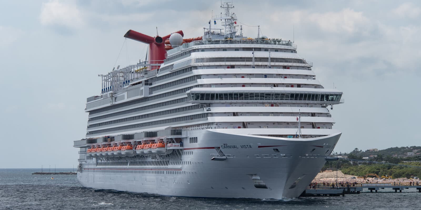 Carnival Vista cruise ship docked in Willemstad harbor, Curaçao, with the ship's red, white, and blue funnel visible against a partly cloudy sky