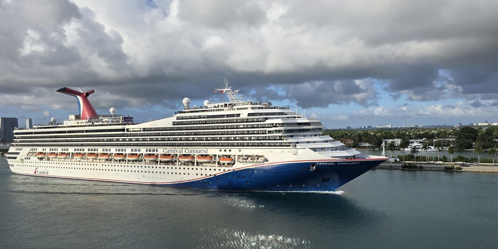 Carnival Conquest cruise ship in Miami waters with Carnival's red, white, and blue whale-tail funnel visible against a partly cloudy sky.