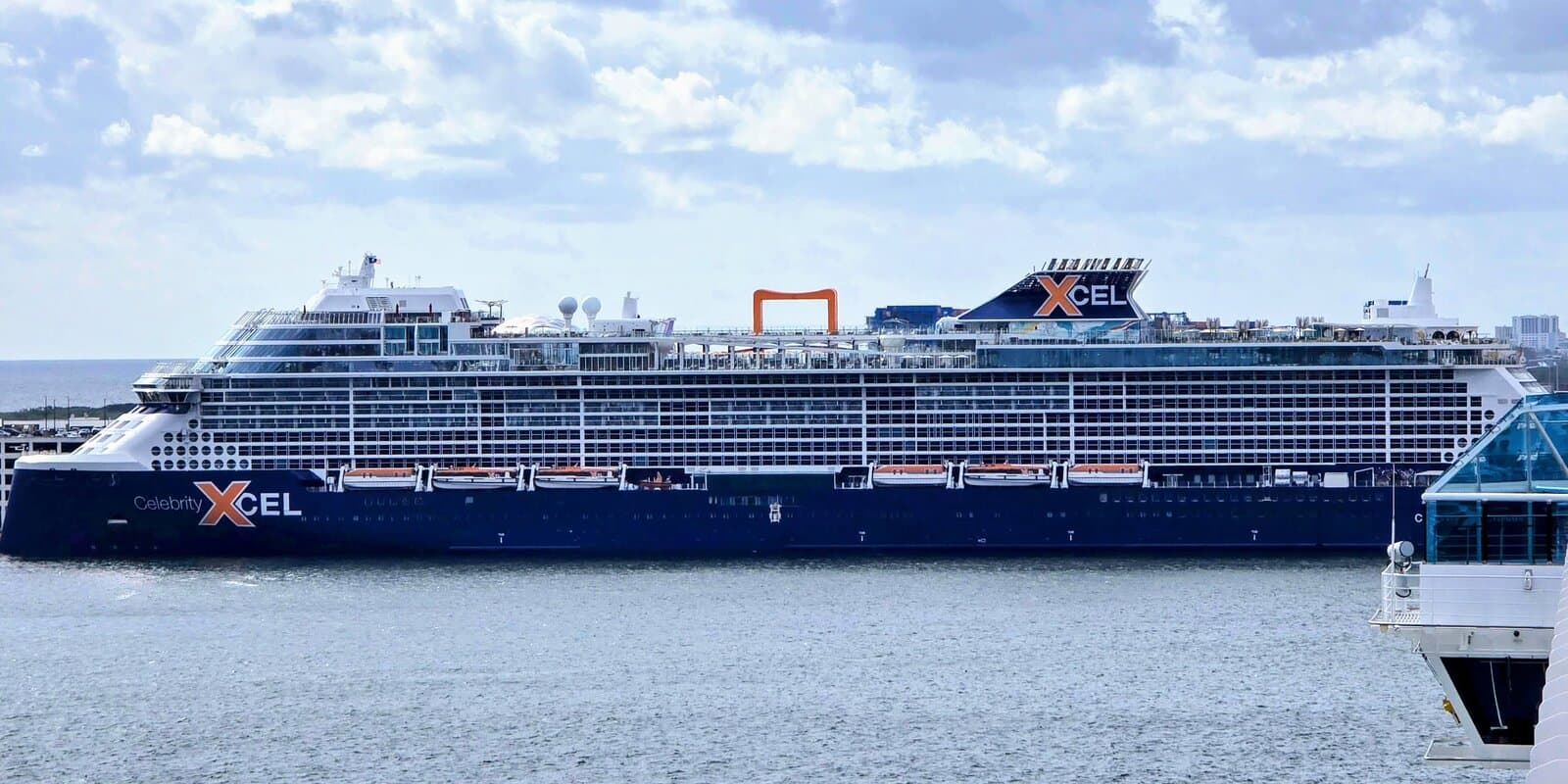 Celebrity Xcel cruise ship at port showing Edge-class hull with orange Magic Carpet on the starboard side and XCEL name on the bow