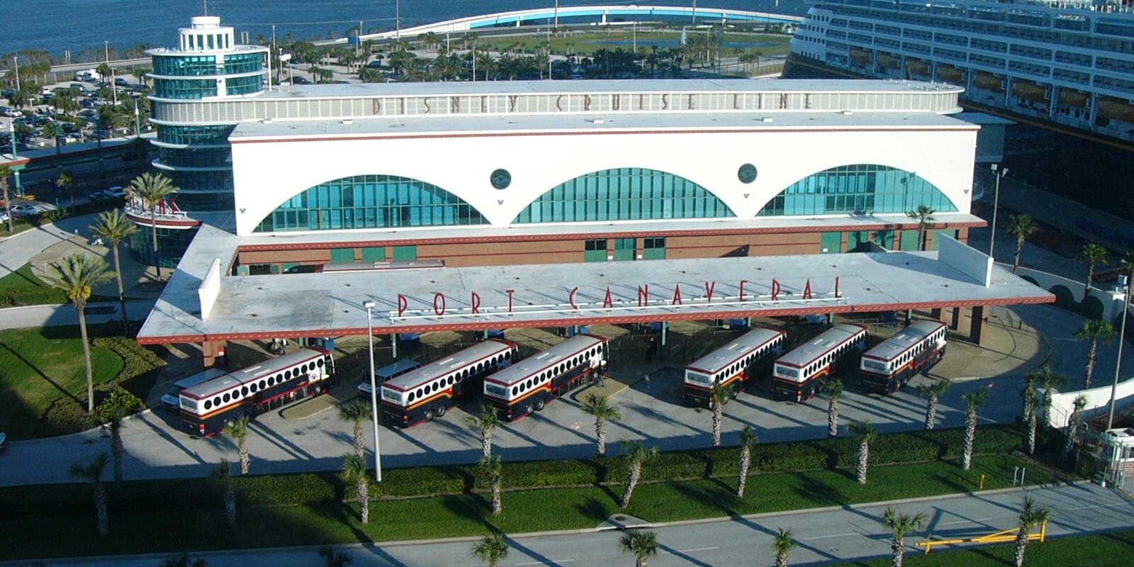 Disney Cruise Line terminal at Port Canaveral with a cruise ship docked alongside and shuttle buses under the passenger drop-off canopy