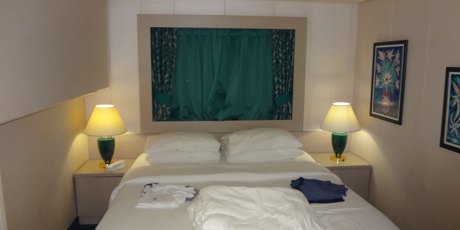 Typical cruise ship stateroom interior with twin beds, window with teal curtains, and bedside lamps