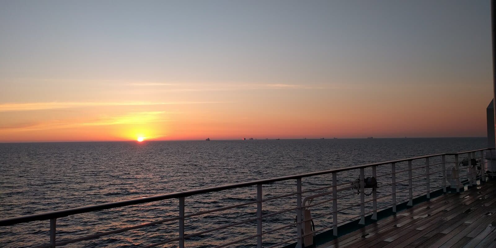 View from a cruise ship deck railing across calm open water toward a sunset horizon