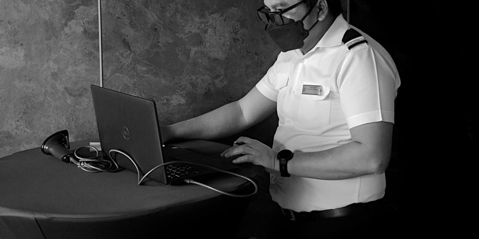A cruise ship crew member operates a card scanner at a laptop during onboard checkout at a payment terminal