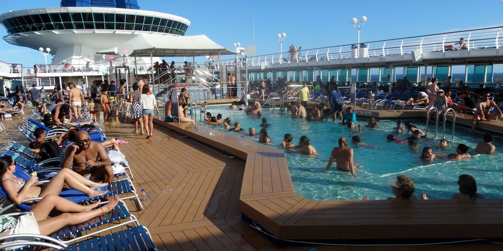 Passengers swimming in a cruise ship pool on a sunny day, with blue-and-white striped lounge chairs on the surrounding deck