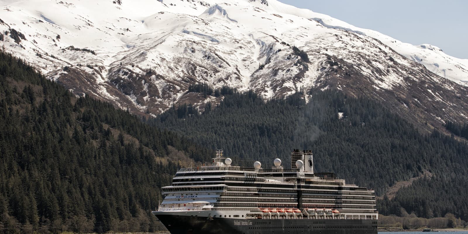 Holland America Line cruise ship Eurodam on calm Alaska waters with snow-capped mountains and evergreen forest along the shore
