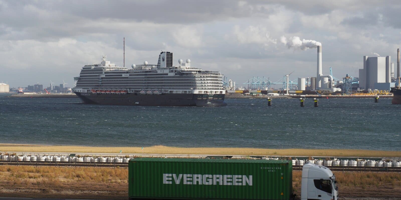 Holland America Line MS Rotterdam in profile at Rotterdam's Maasvlakte port, navy hull and white superstructure with Holland America branding, container cranes in the background.