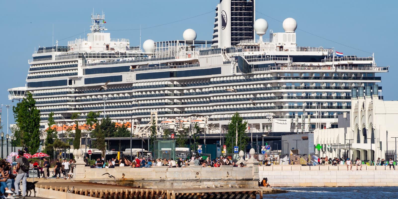 MS Nieuw Statendam docked at a Lisbon pier on the Tagus River, white hull and HAL funnel visible with passengers on the waterfront promenade