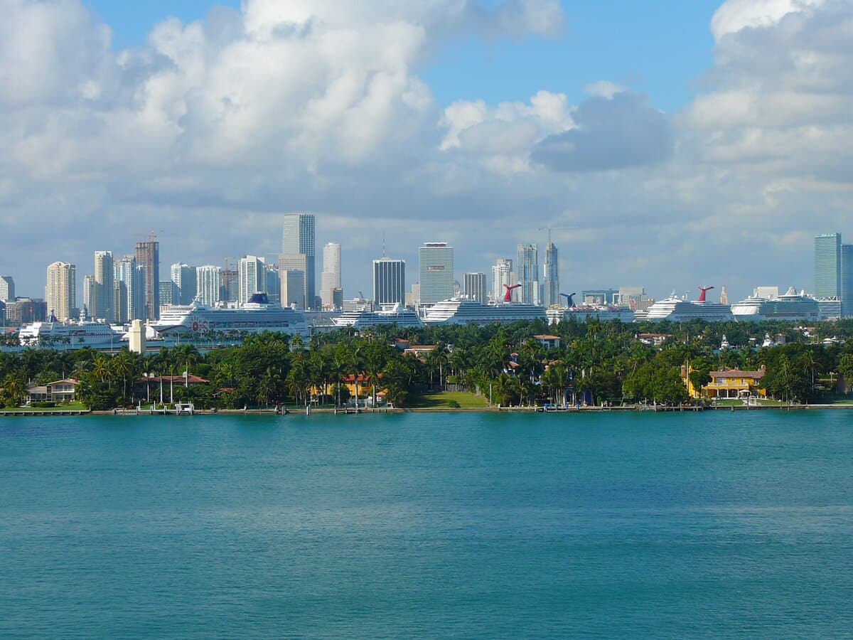 Seven cruise ships docked at PortMiami with downtown Miami skyscrapers and Biscayne Bay in the foreground