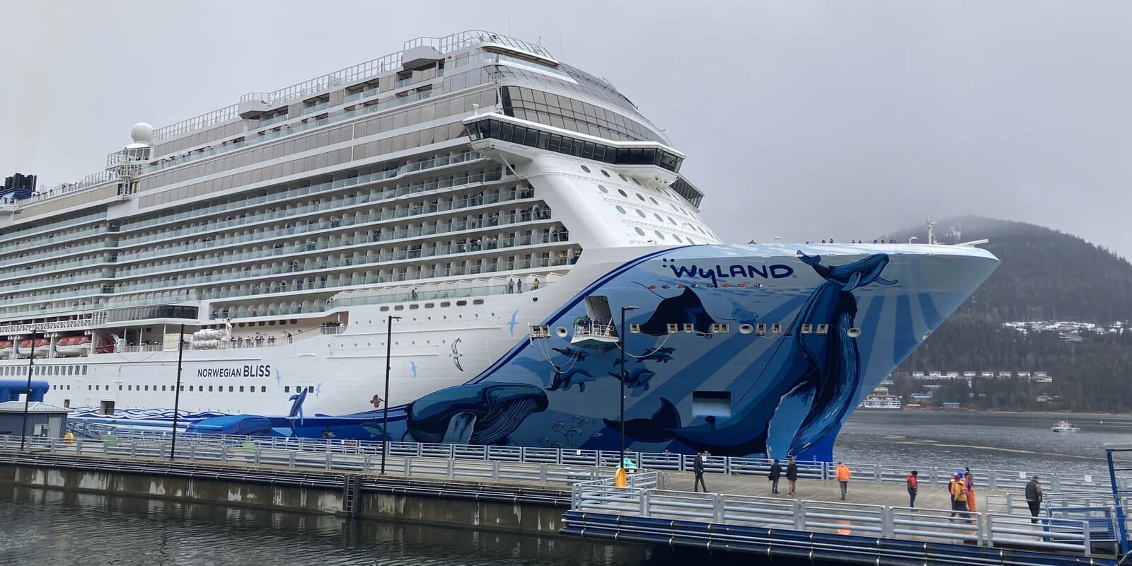 Norwegian Bliss docked at a pier in Juneau, Alaska, with forested mountains under overcast skies