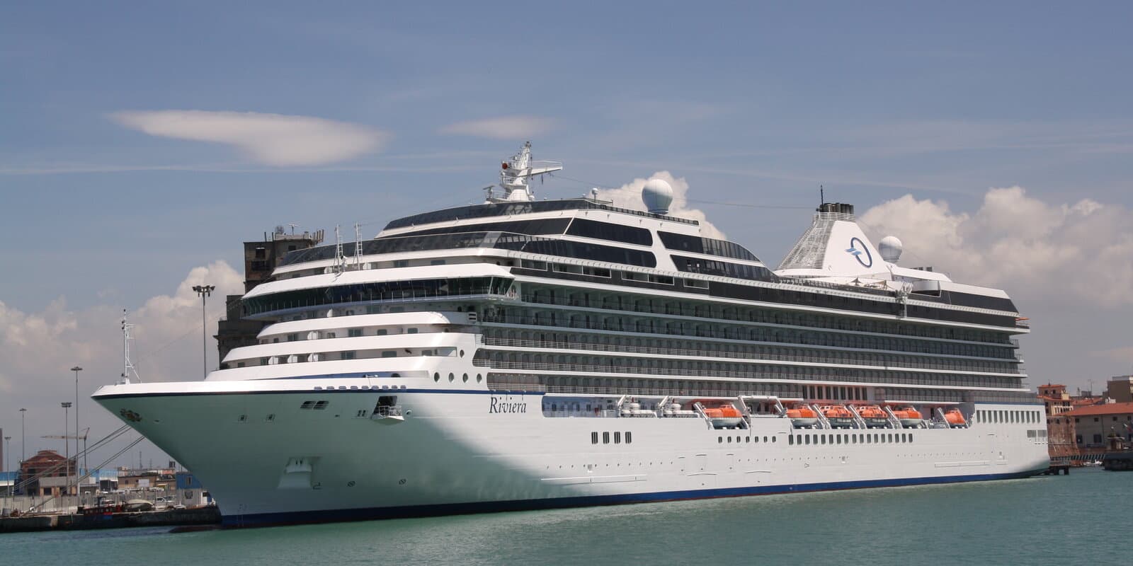 Oceania Riviera cruise ship docked at a Mediterranean port with turquoise water and terracotta-roof buildings in the background
