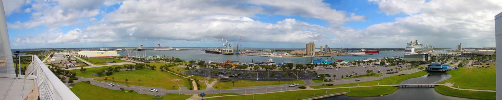 Panoramic view of Port Canaveral with Carnival and Royal Caribbean cruise ships at terminals, cargo cranes, and the port basin under a partly cloudy sky