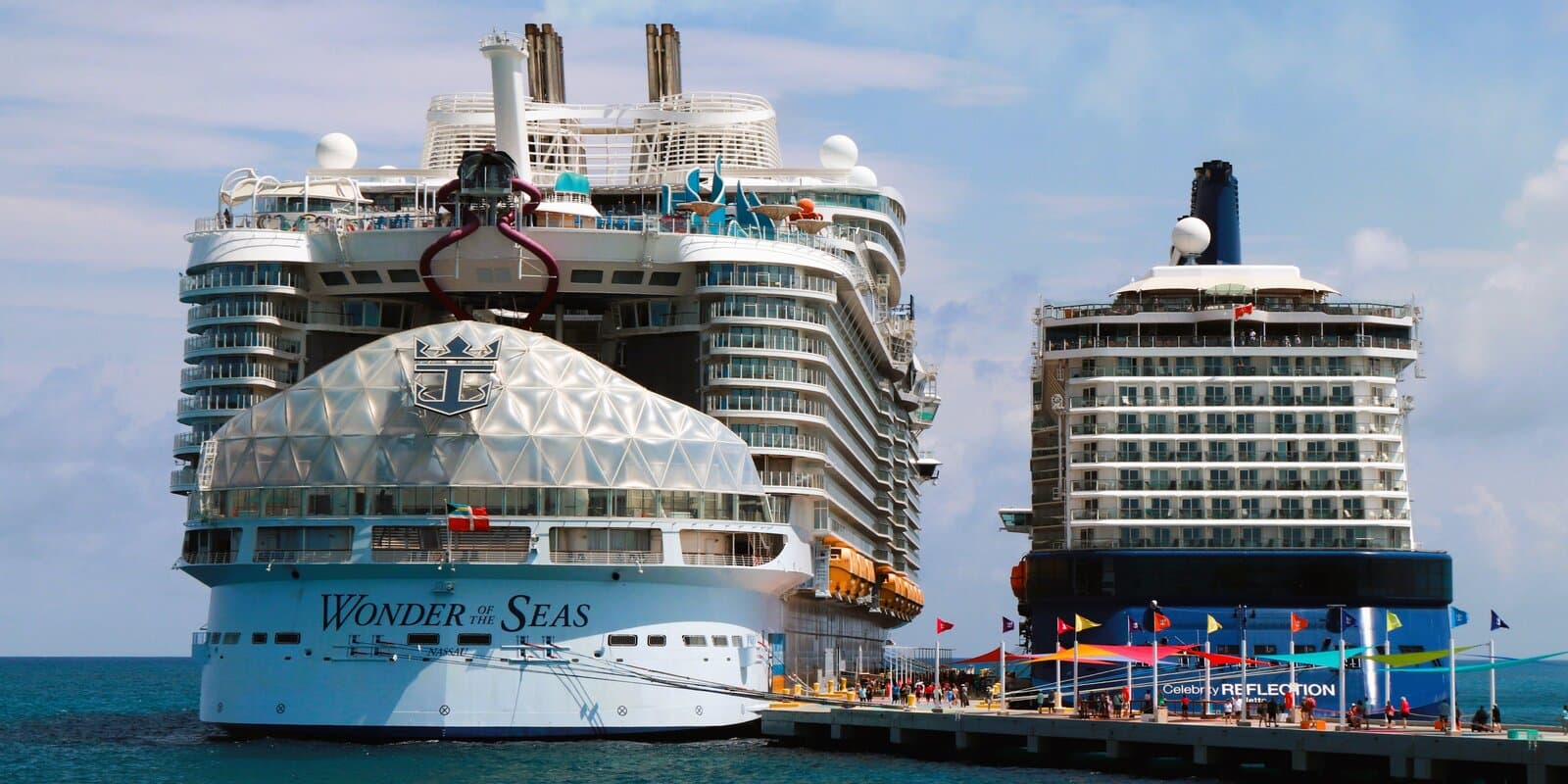 Wonder of the Seas and Celebrity Reflection cruise ships docked stern-to-stern at Perfect Day at CocoCay in the Bahamas, with turquoise water and a pier walkway between them