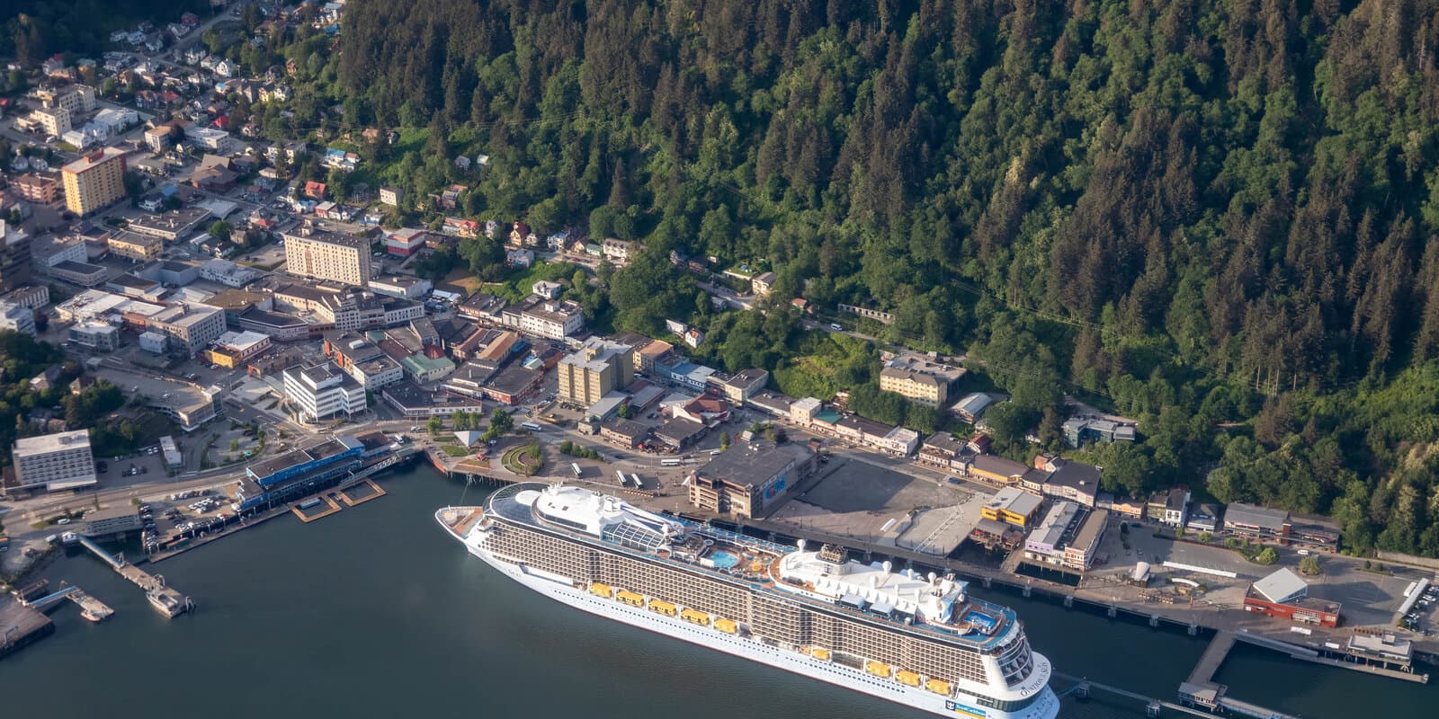 Aerial view of Ovation of the Seas docked at a pier in Juneau, Alaska, with forested mountains rising behind the waterfront town