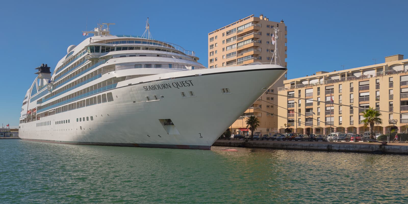 Seabourn Quest moored bow-in at a Mediterranean port with the ship name on the hull, waterfront buildings, and palm trees along the pier