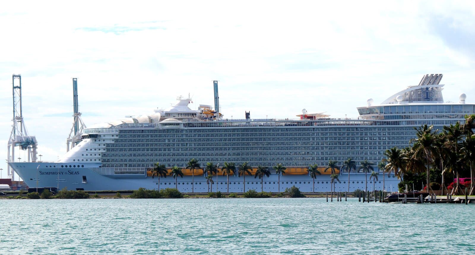 Symphony of the Seas docked at PortMiami with the ship name on the bow, yellow lifeboats along the hull, and the Royal Caribbean funnel visible against a bright sky