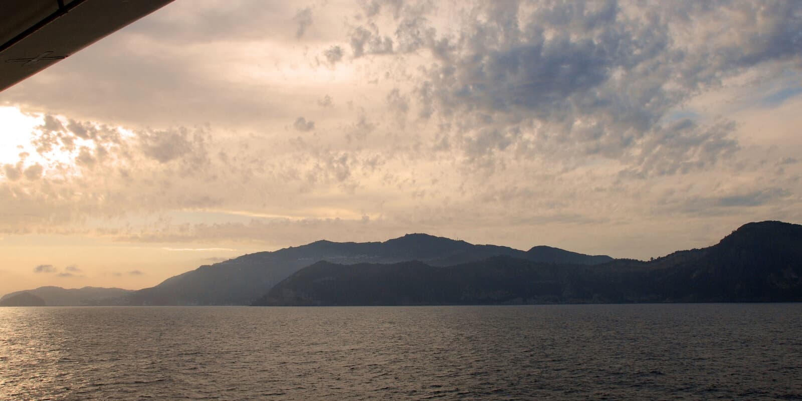 View from a cruise ship deck toward a mountainous Mediterranean coastline at dusk with calm water