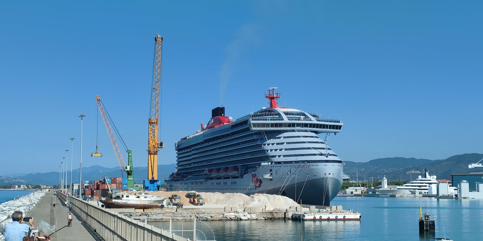 Valiant Lady cruise ship docked at Marina di Carrara port with yellow cranes, gravel piles on the pier, and VALIANT LADY lettering on the grey hull