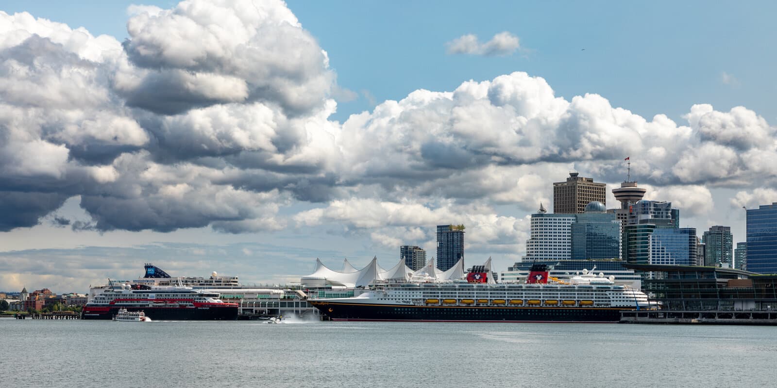 Cruise ships docked at Canada Place in downtown Vancouver, with the terminal's white sail-shaped roof, the Vancouver skyline, and Burrard Inlet in the foreground