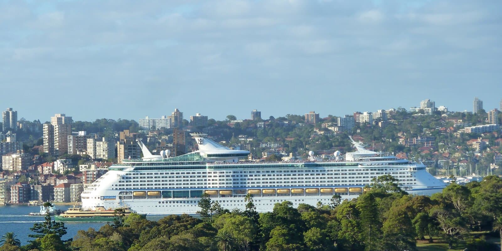 Royal Caribbean Voyager of the Seas with hull name visible, sailing in harbor with city buildings beyond