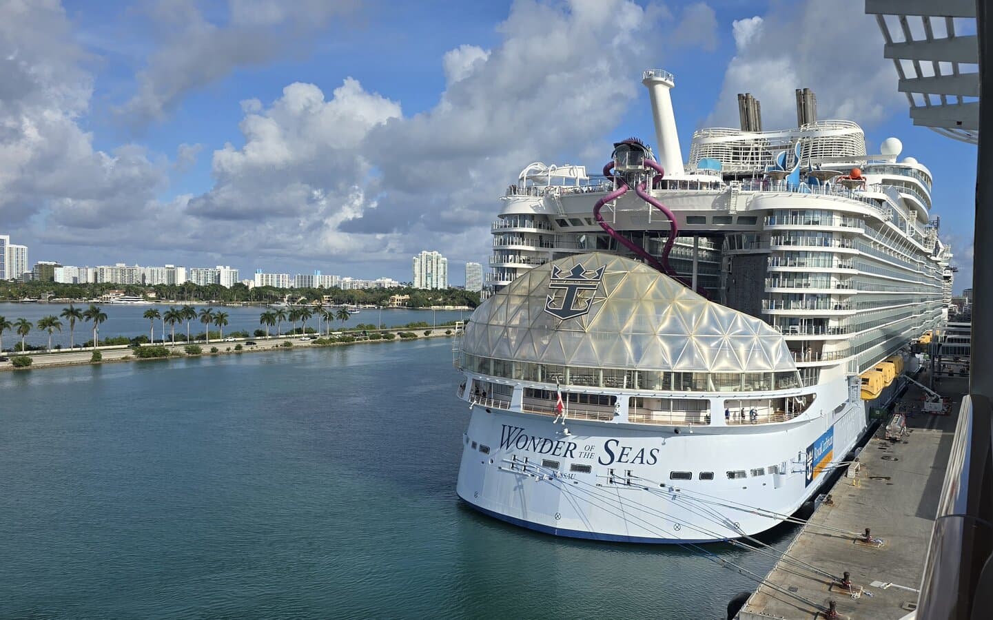 Wonder of the Seas docked at PortMiami with the ship name on the stern, AquaTheater dome, and Ultimate Abyss slides visible against a blue sky
