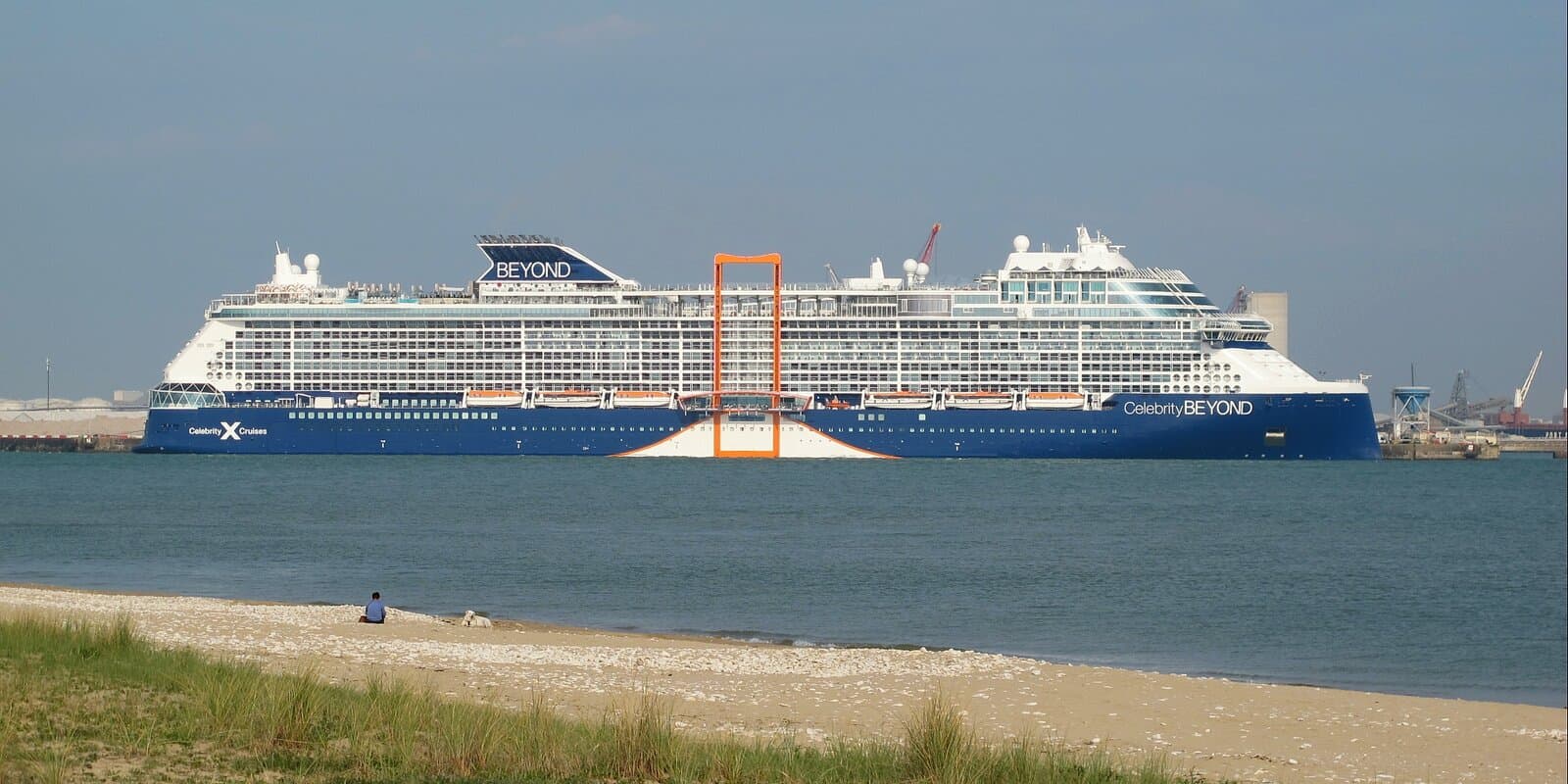 Celebrity Beyond cruise ship with navy hull and white decks sailing past a sandy beach, Celebrity branding and orange Magic Carpet visible