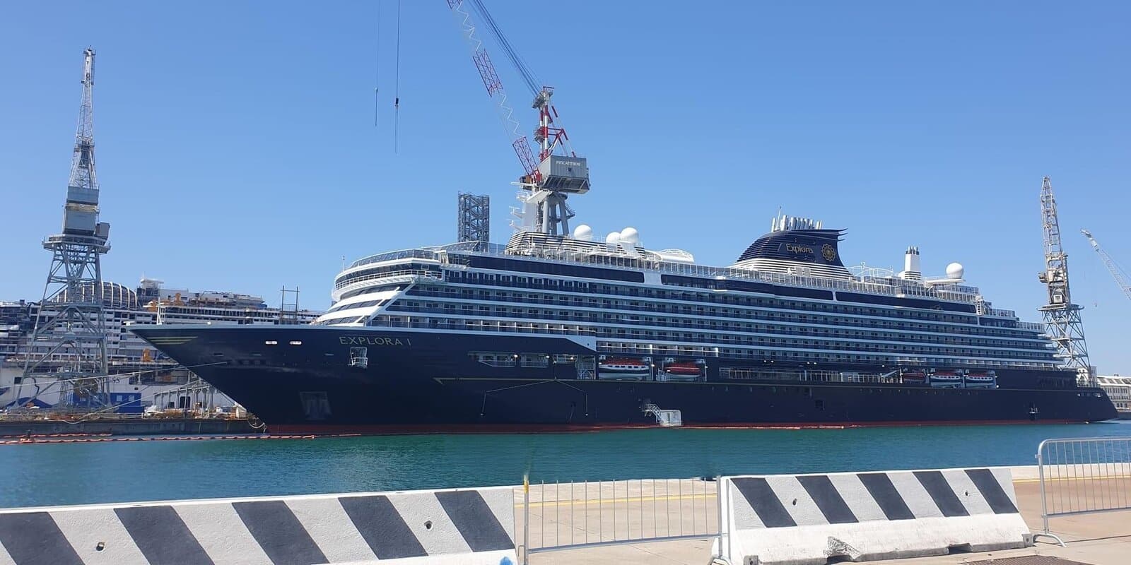 Explora I, a sister ship to Explora III, dark hull and white decks docked at a shipyard pier under a clear blue sky