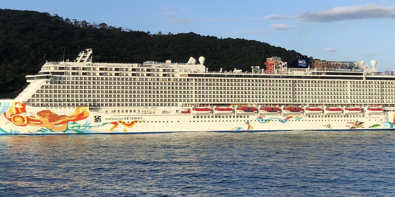 Norwegian Getaway cruise ship in starboard profile on blue water with a green hillside behind, colorful hull art and NCL funnel visible