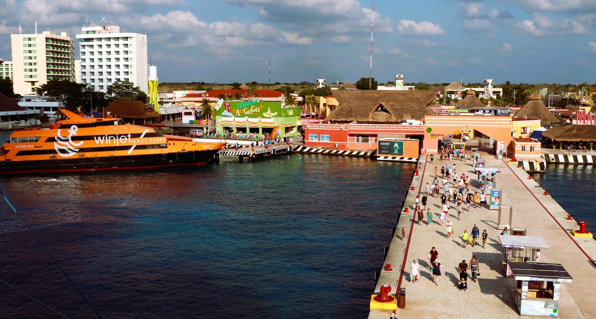 Passengers walking along the Cozumel cruise pier past waterfront shops and a docked ferry