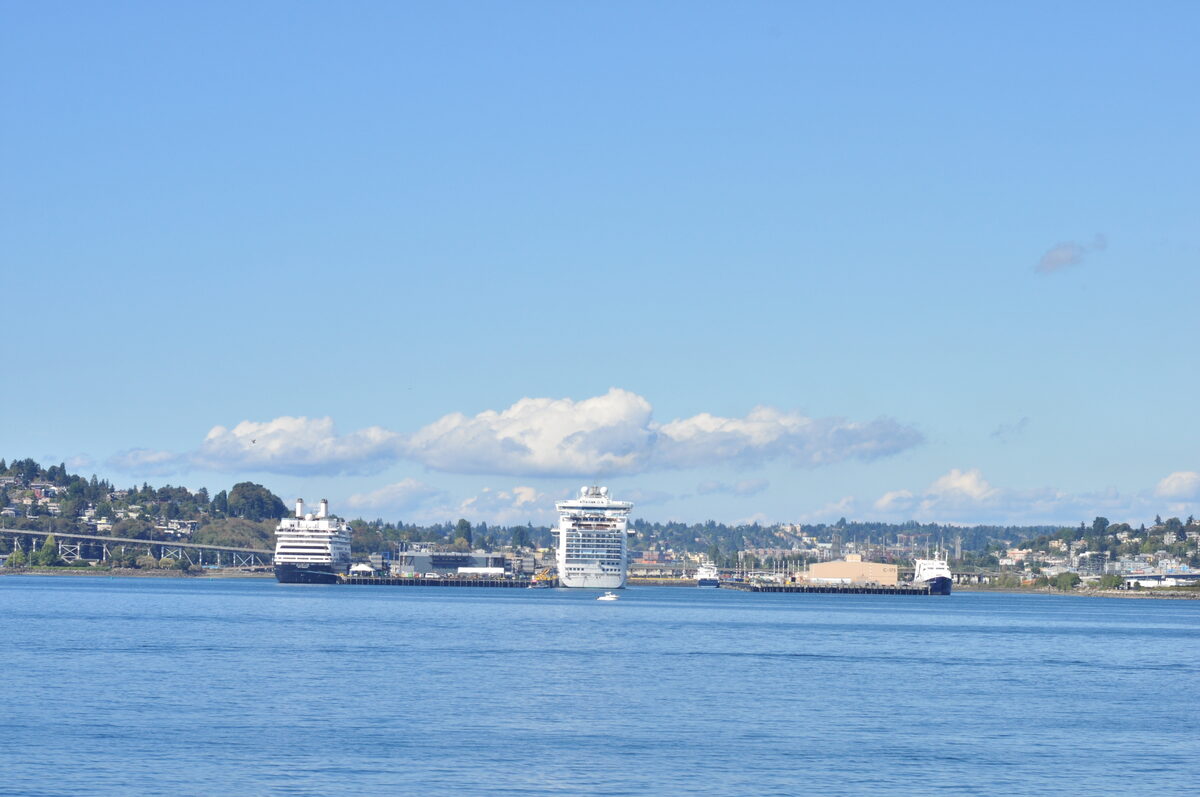 Holland America Amsterdam and Ruby Princess docked at Smith Cove piers 90–91 in Seattle, with downtown Seattle and Queen Anne Hill visible across Elliott Bay