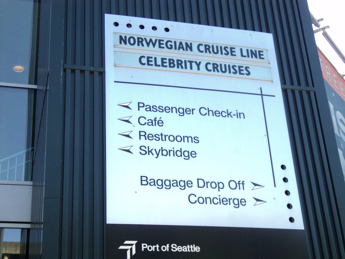 Vertical wayfinding sign at a cruise terminal listing passenger check-in, baggage drop-off, and concierge directions