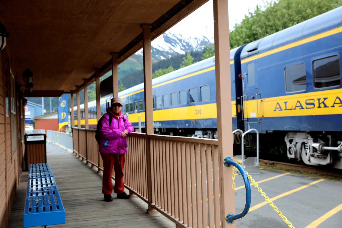 Traveler on the Alaska Railroad platform beside a blue and yellow train car at the Seward depot, with snow-capped mountains behind