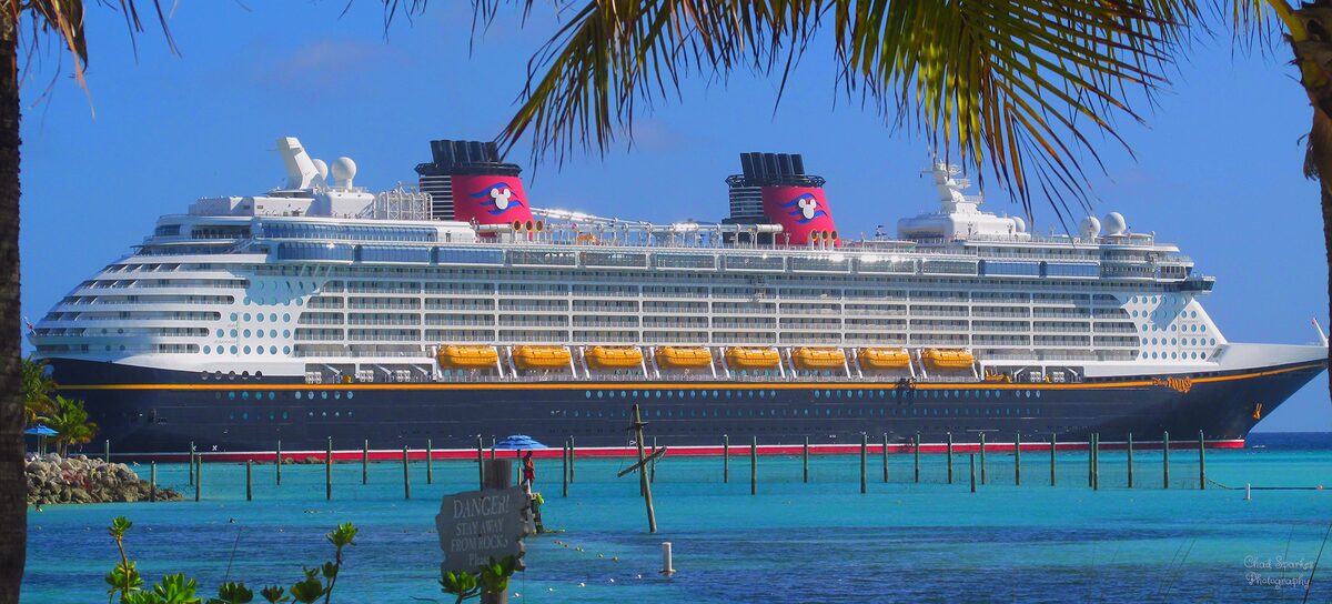 Disney Fantasy cruise ship docked at Castaway Cay in the Bahamas, with the ship's colorful funnel visible against a blue sky and calm turquoise water