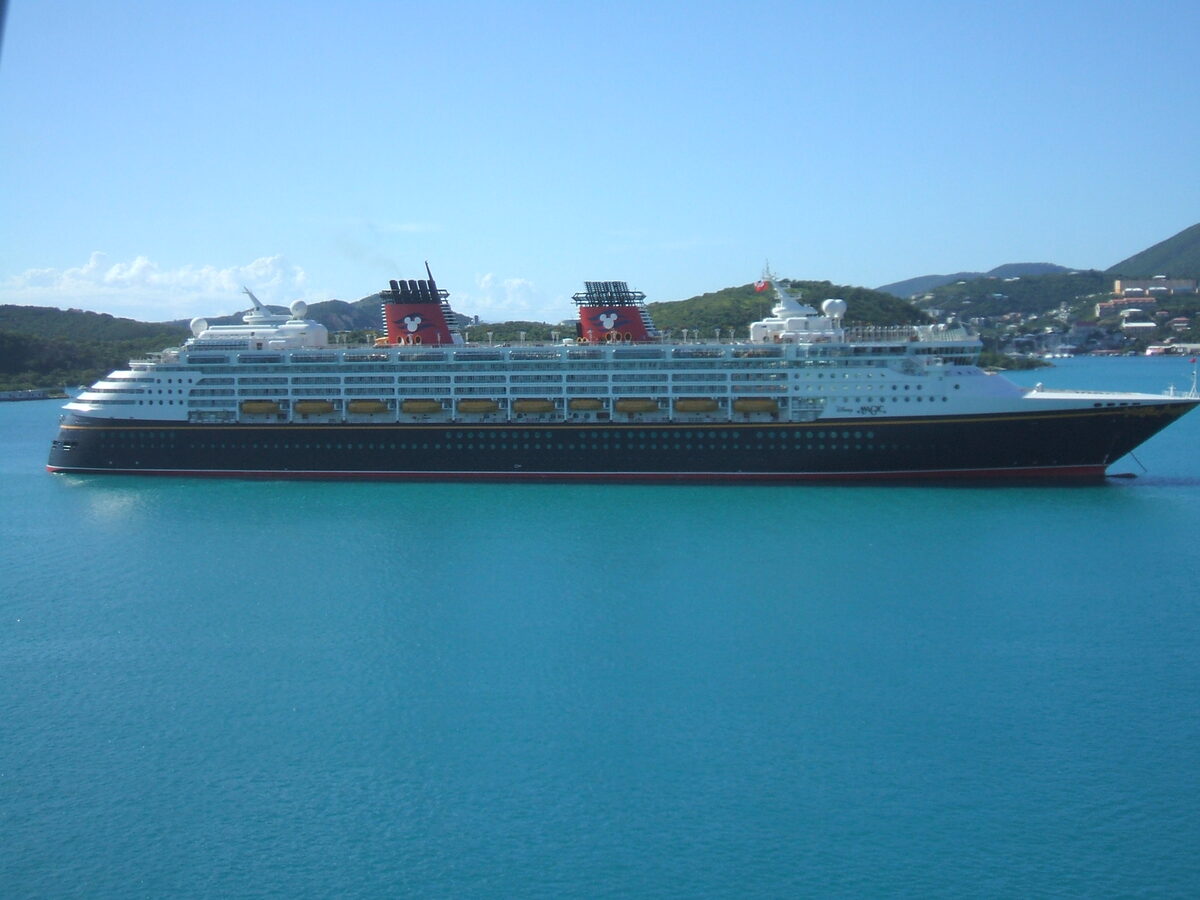 Disney Magic cruise ship at anchor on calm turquoise water, with Mickey Mouse logos on its red funnels