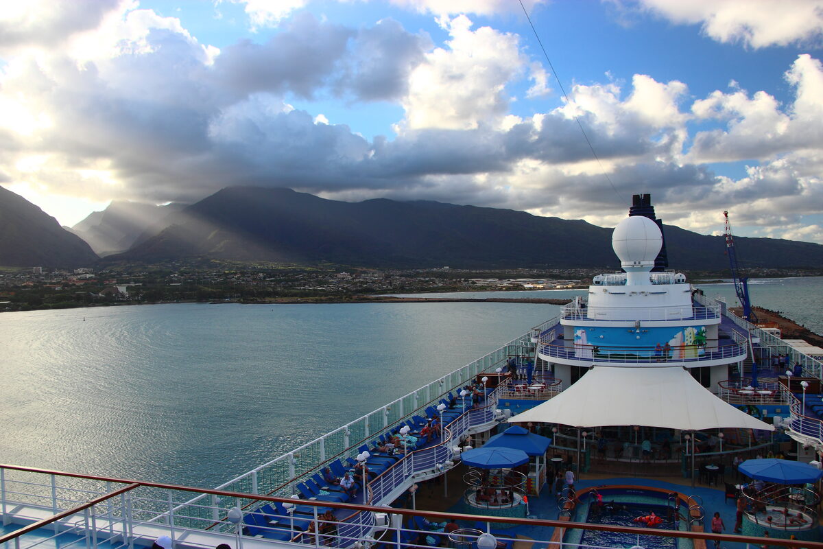 View from Pride of America's upper deck showing the ship docked in Hilo Bay with volcanic mountains and sun rays through clouds