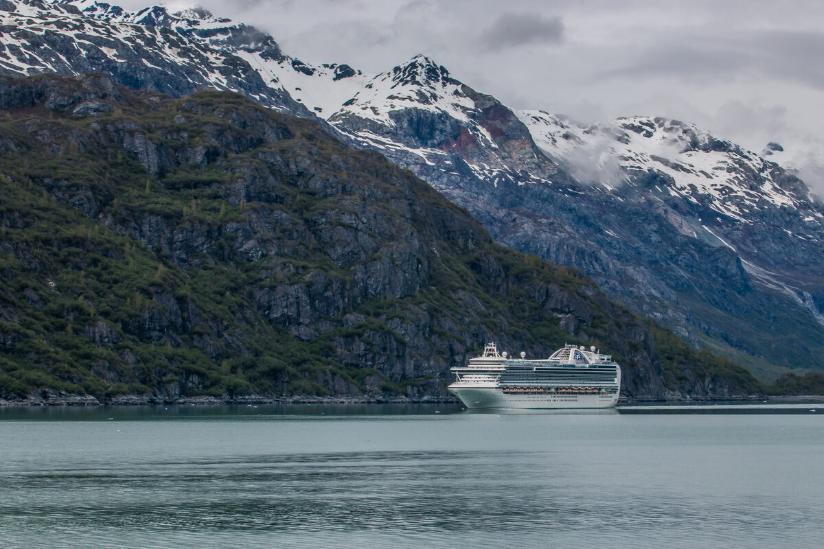 A cruise ship sails past steep, snow-dusted mountains along Alaska's Inside Passage on an overcast day