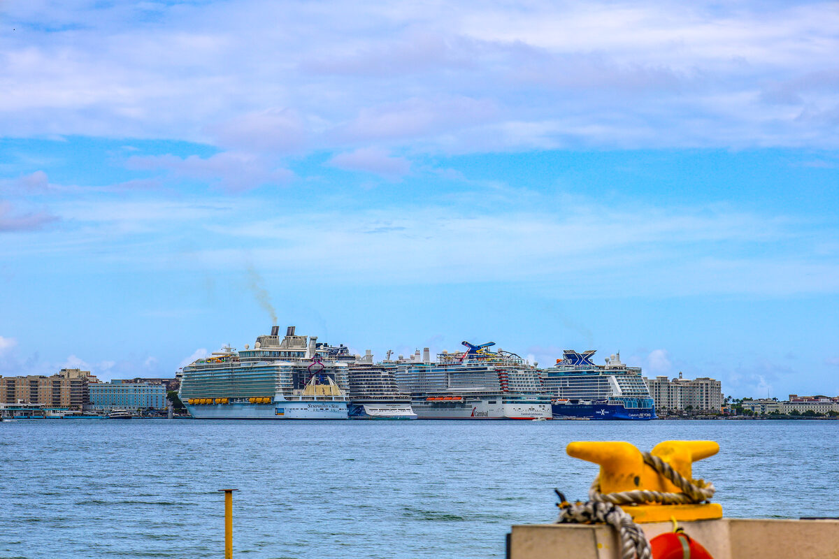 Four cruise ships docked side by side at the Old San Juan cruise port in Puerto Rico, viewed across the harbor with a yellow mooring bollard in the foreground