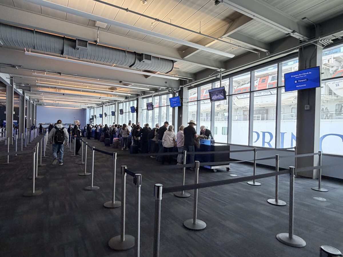 Passengers line up at embarkation check-in counters in a cruise terminal with a docked ship visible through floor-to-ceiling windows