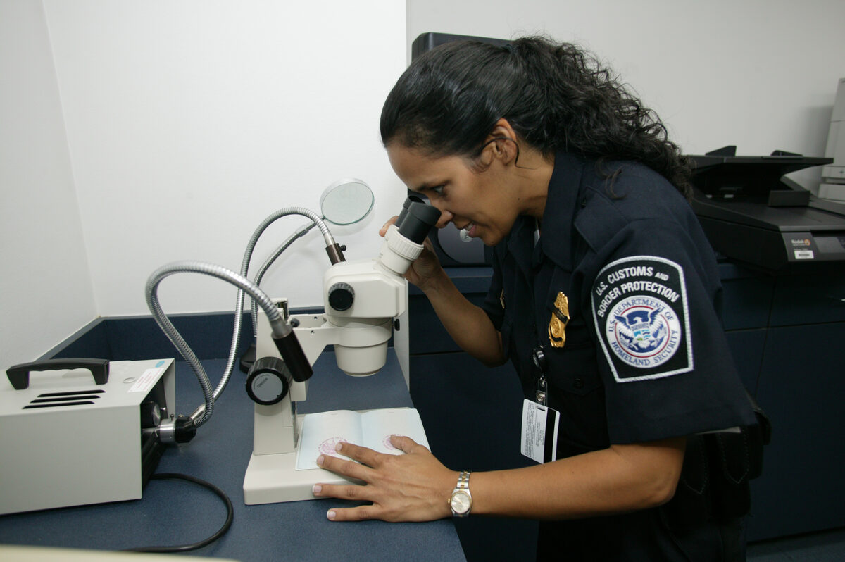 A customs officer examines an open passport under magnification at a document verification desk